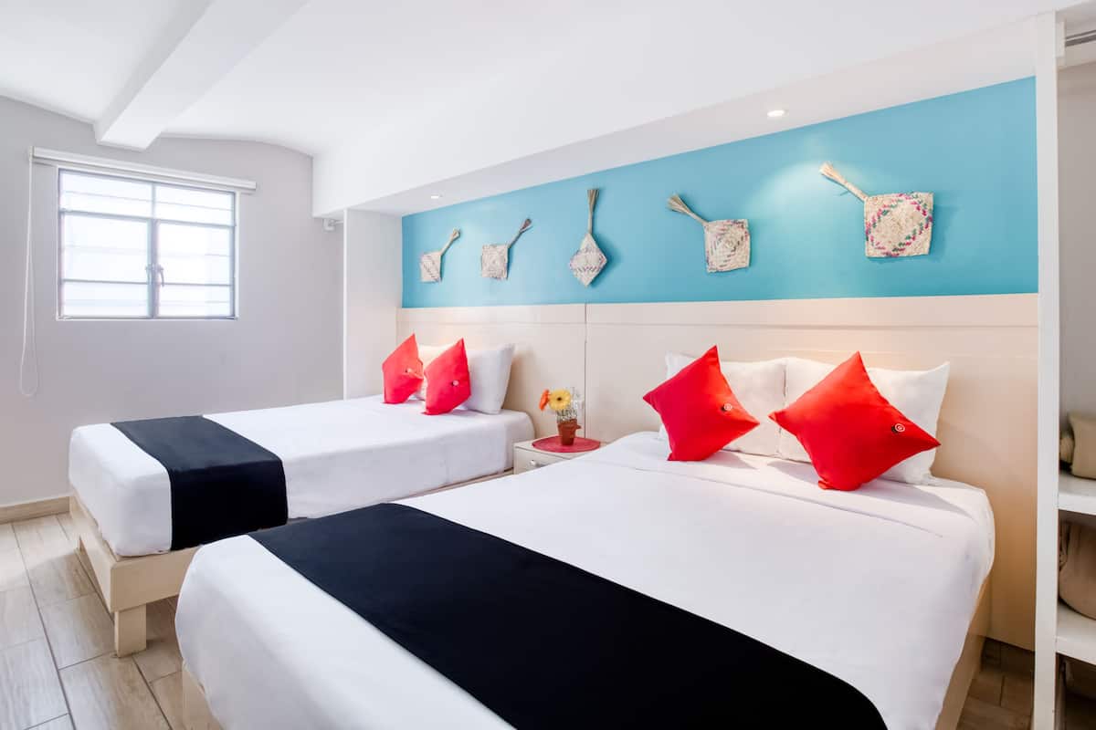 A hotel room with two beds, each with white bedding and black and red pillows. The room has a blue accent wall and a window with blinds.