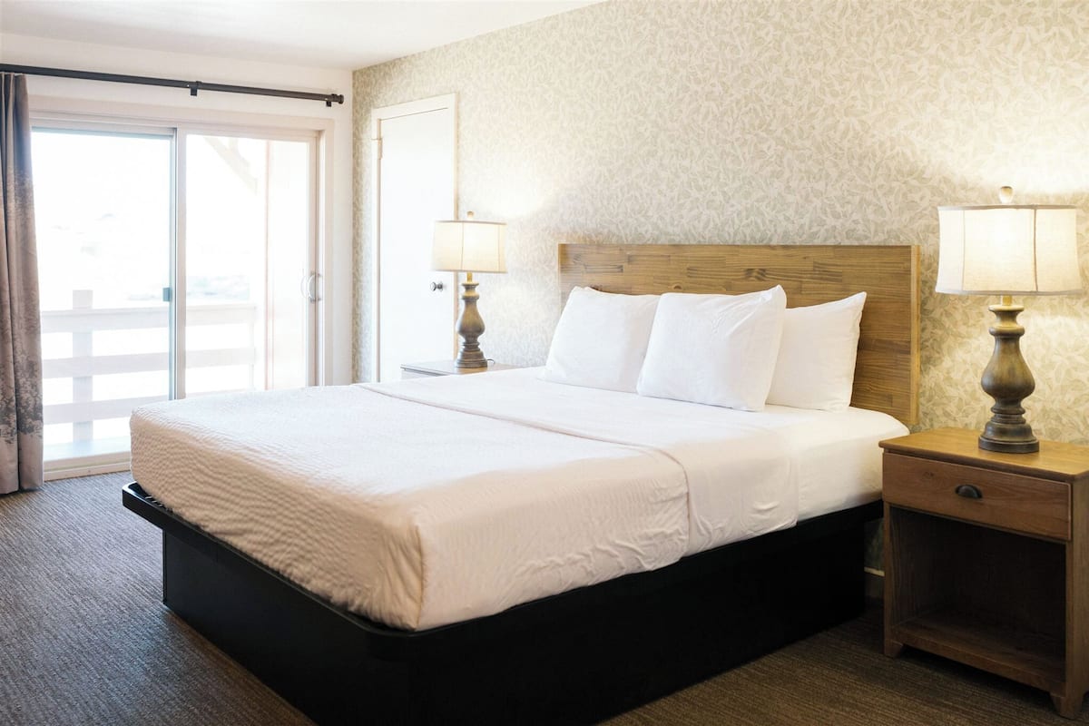 A hotel room with a large bed, two bedside lamps, a wooden headboard, and a sliding glass door.