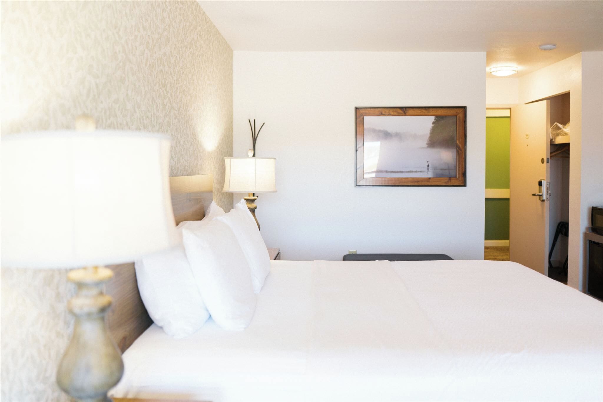 A hotel room with a bed, a lamp, a framed picture on the wall, and a door leading to another room.