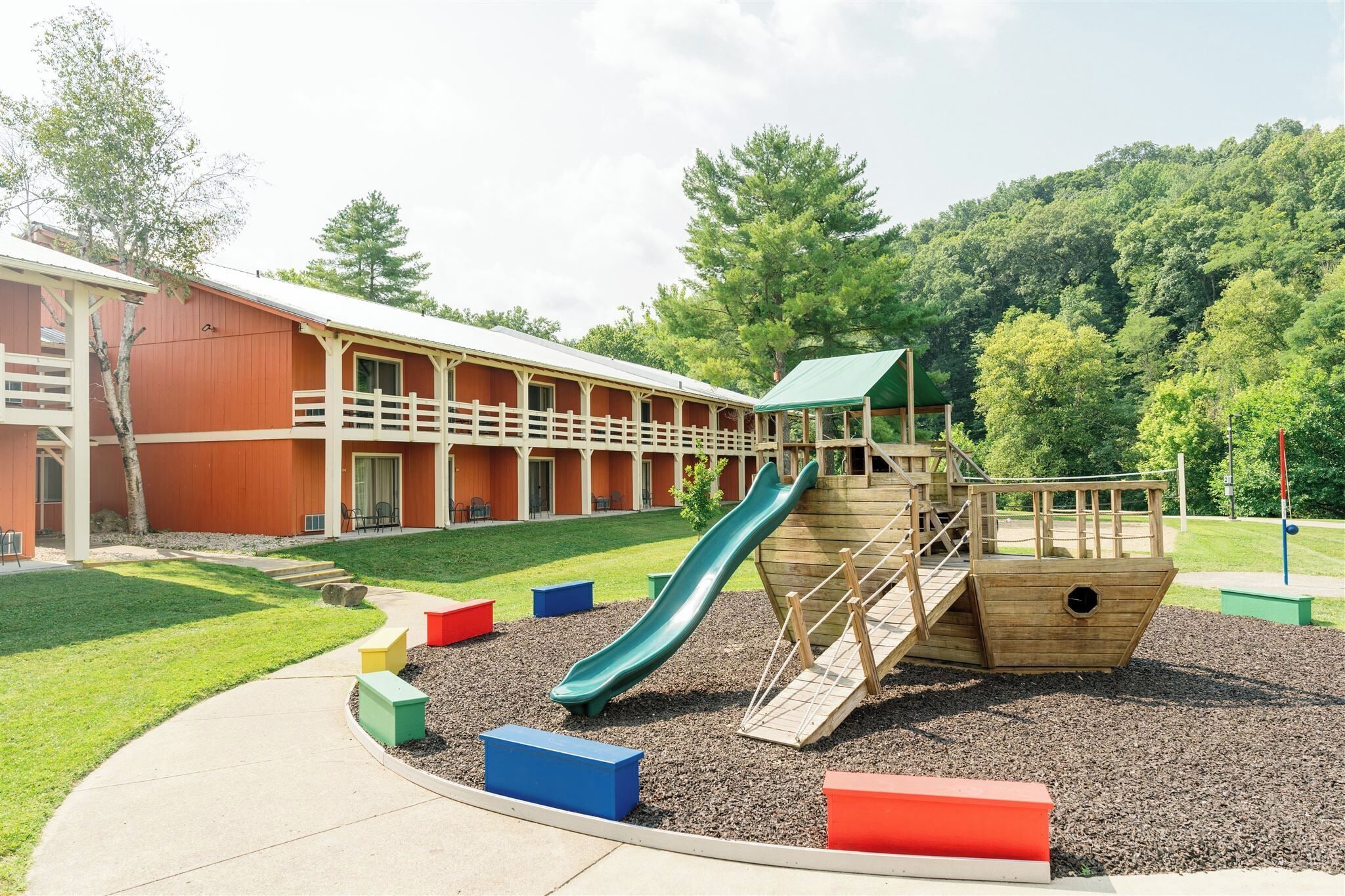 A playground with a slide and climbing structures, surrounded by a building with a red exterior and a covered walkway.
