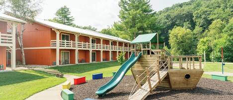 A playground with a slide and climbing structures, surrounded by a building with a red exterior and a covered walkway.