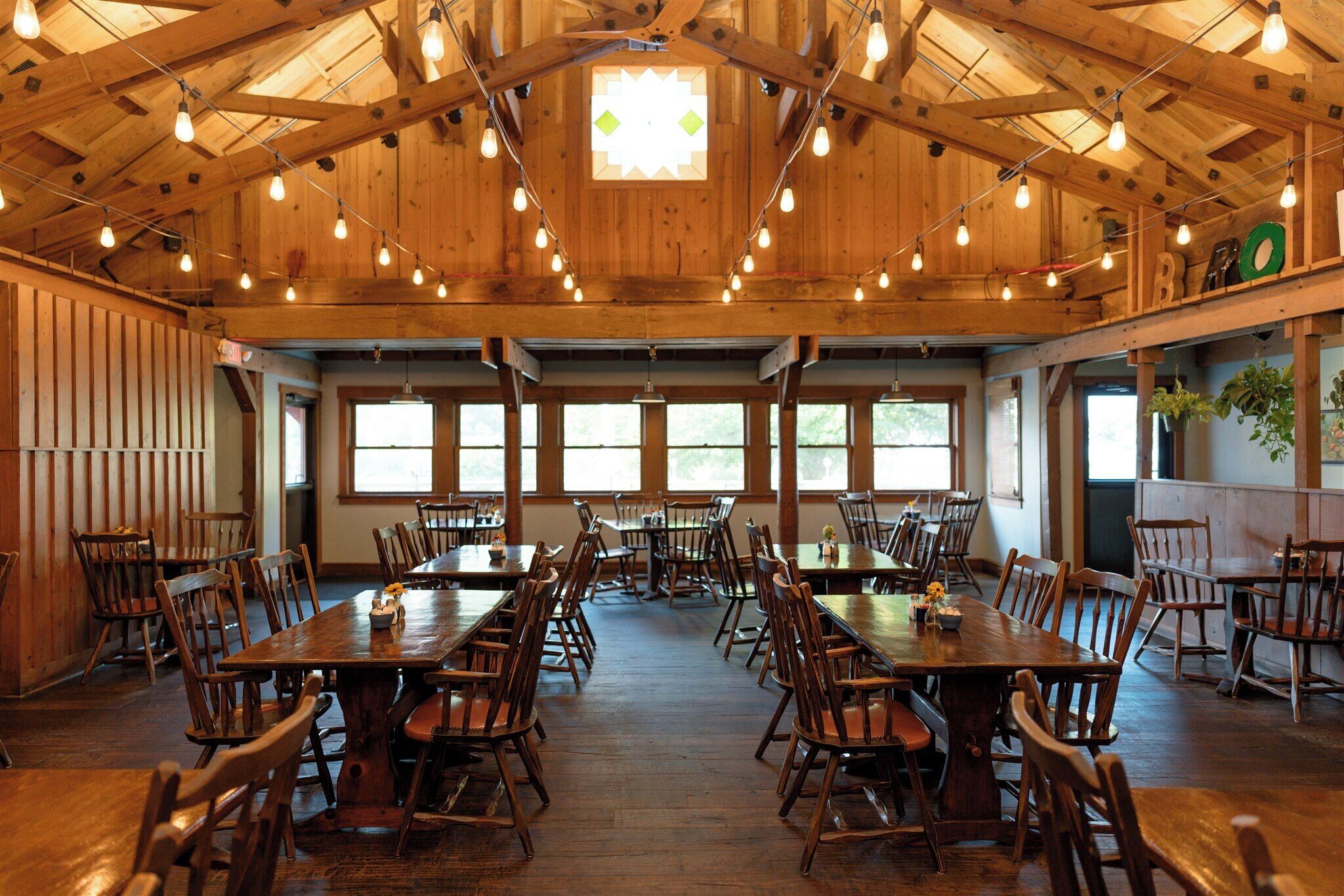 A spacious dining hall with wooden tables and chairs, large windows, and a high ceiling with exposed beams and hanging lights.