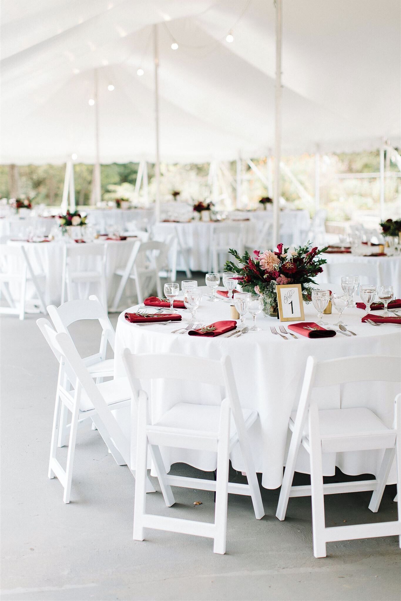 Un espace événementiel extérieur aménagé sous une tente, avec des tables rondes, des chaises blanches et des décorations florales au centre.