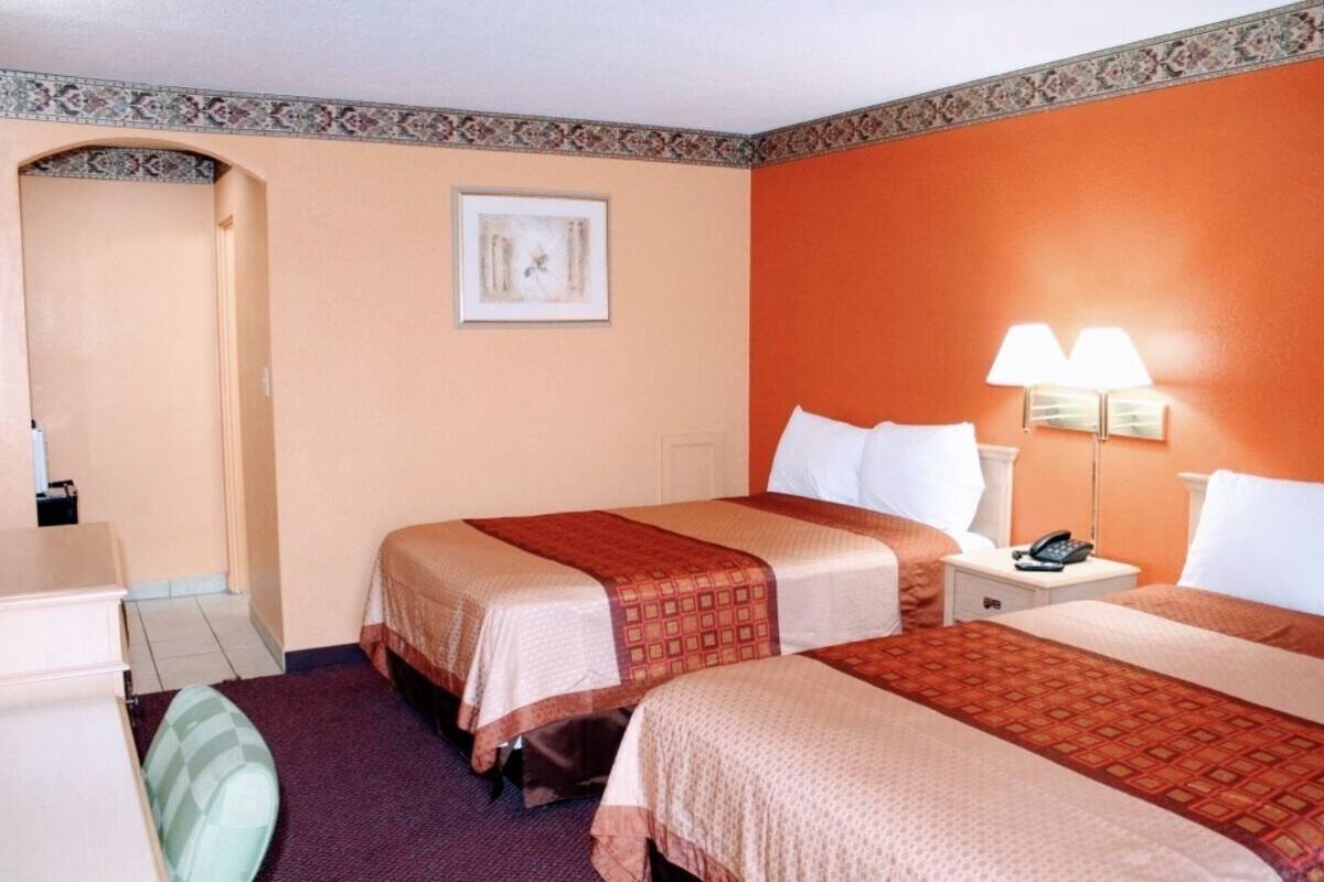 A hotel room with two beds, a desk, and a chair. There is a framed picture on the wall and a lamp on the nightstand.