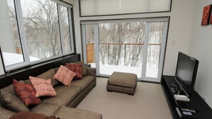 A modern living room with a sectional sofa, a small ottoman, and a flat-screen TV on a black stand. Large windows provide a view of a snowy landscape. - Neyuki (Kutchan)