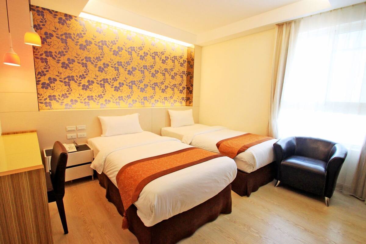 Habitación de hotel con dos camas, un escritorio, una silla y un ventanal grande.