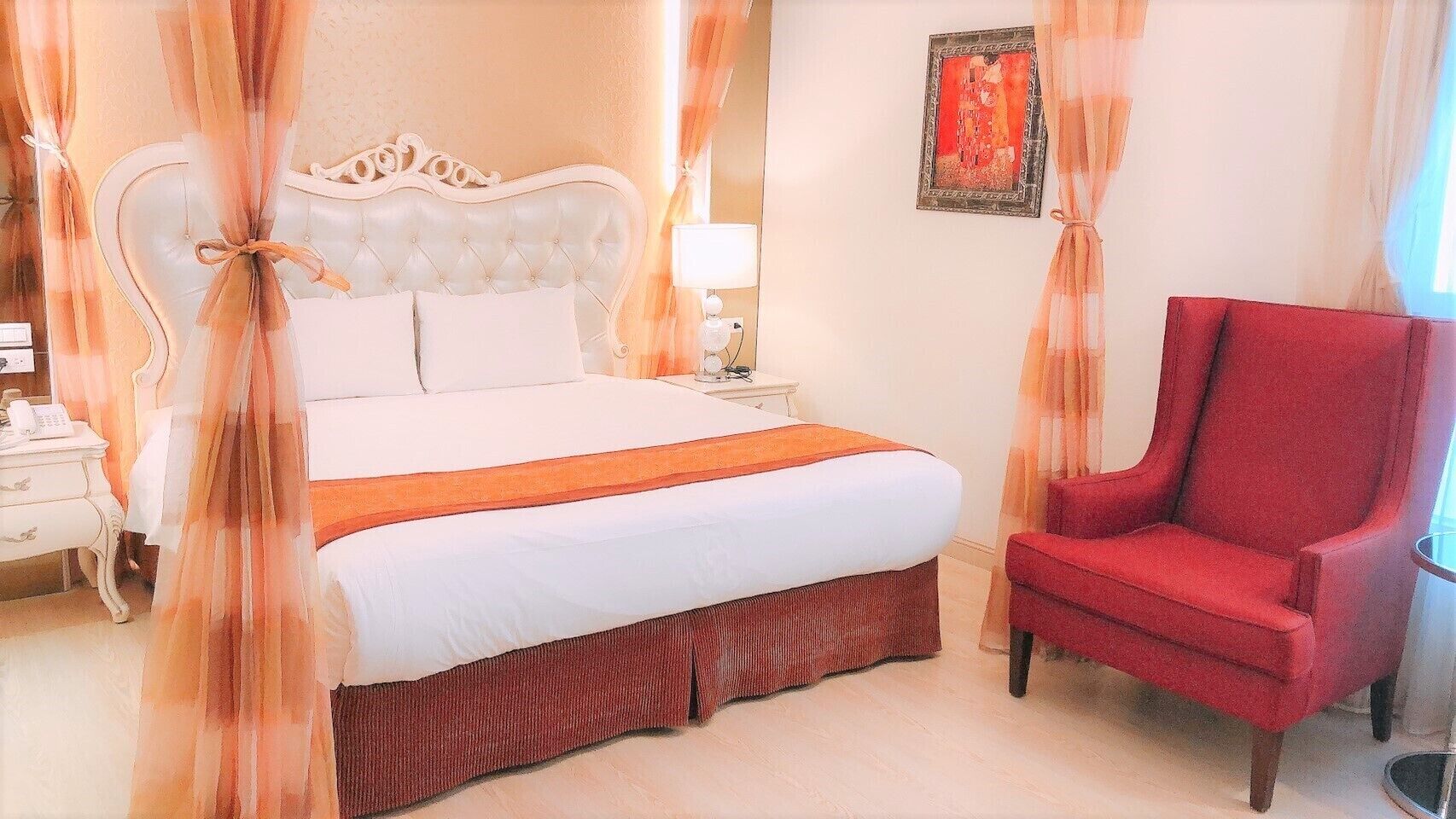 A hotel room with a bed, a red armchair, a nightstand, and a wall-mounted lamp.