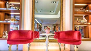 Un hall de hotel con dos sillones tapizados en terciopelo rojo orientados hacia una mesa blanca central, rodeado de estanterías de madera con objetos decorativos.