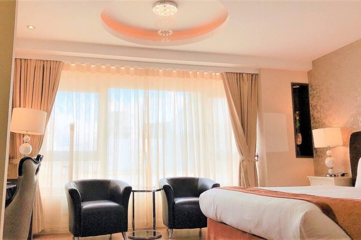 Habitación de hotel con cama, dos sillas, un escritorio y un ventanal grande con cortinas.