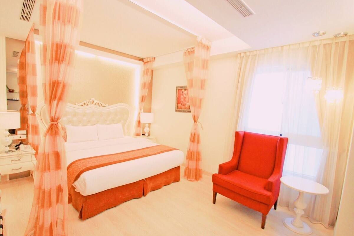 Un dormitorio con una cama grande, un sillón rojo, una mesita y cortinas traslúcidas.