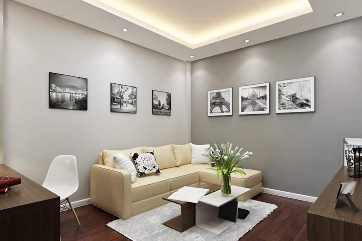 A modern living room with a beige sectional sofa, a white coffee table, and a small side table. There are six black-and-white photographs on the wall.