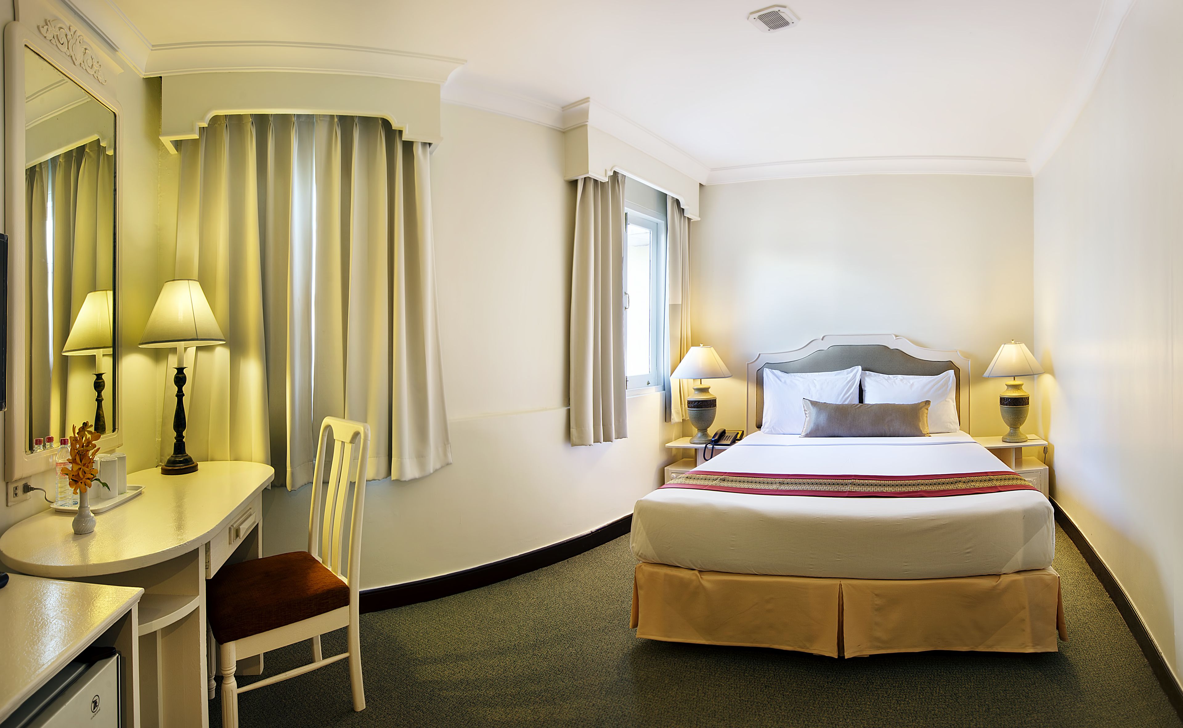 A hotel room with a bed, two bedside lamps, a desk with a chair, a mirror, and curtains.