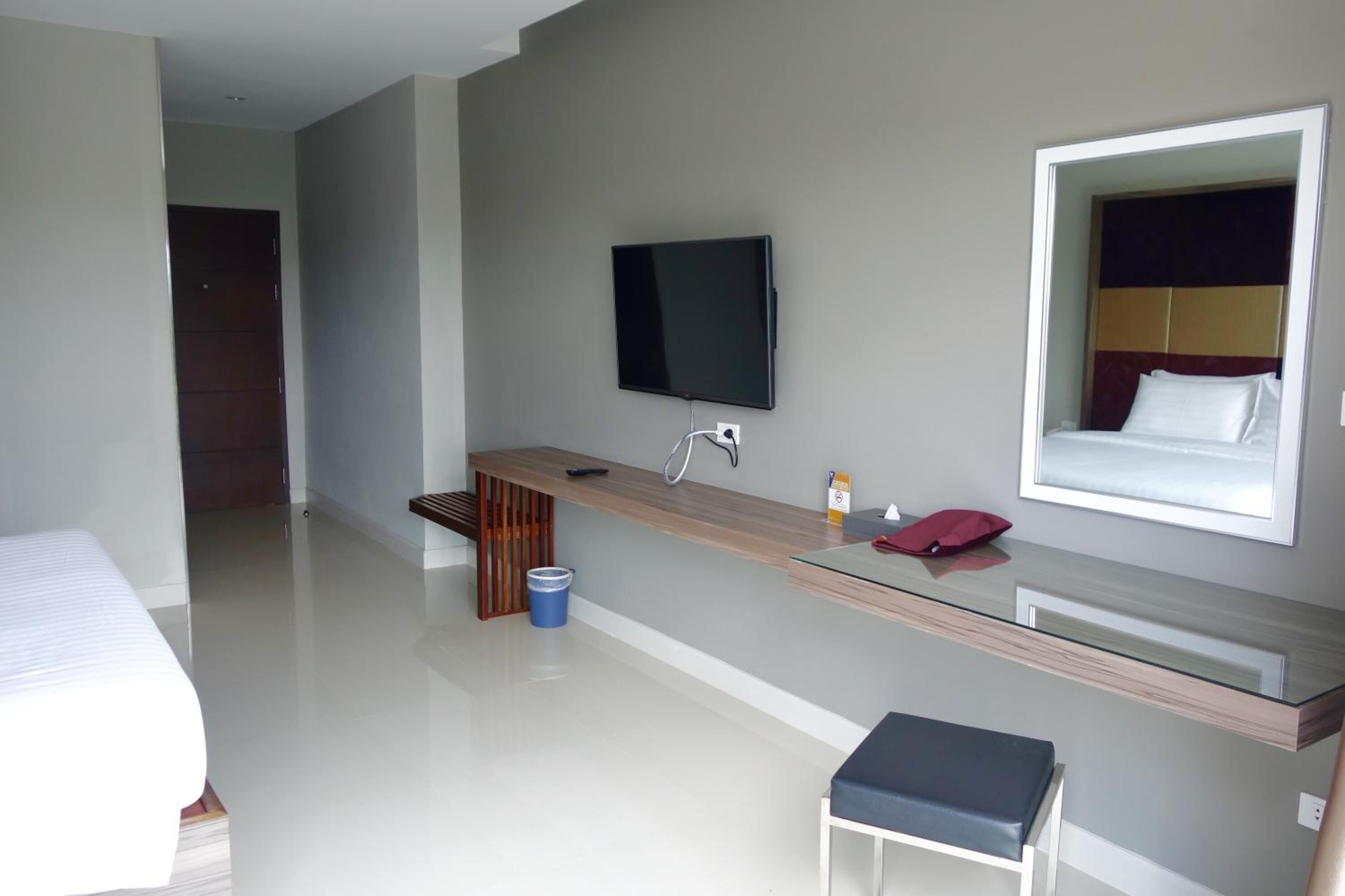 A modern hotel room with a bed, a desk with a flat-screen TV, a mirror, and a small stool.