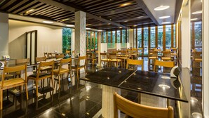 A spacious indoor area with wooden chairs and tables, large windows, and a modern ceiling design.