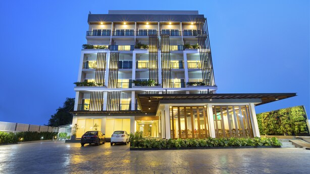 A multi-story hotel with a modern design, featuring balconies with plants, a well-lit entrance, and a paved driveway.
