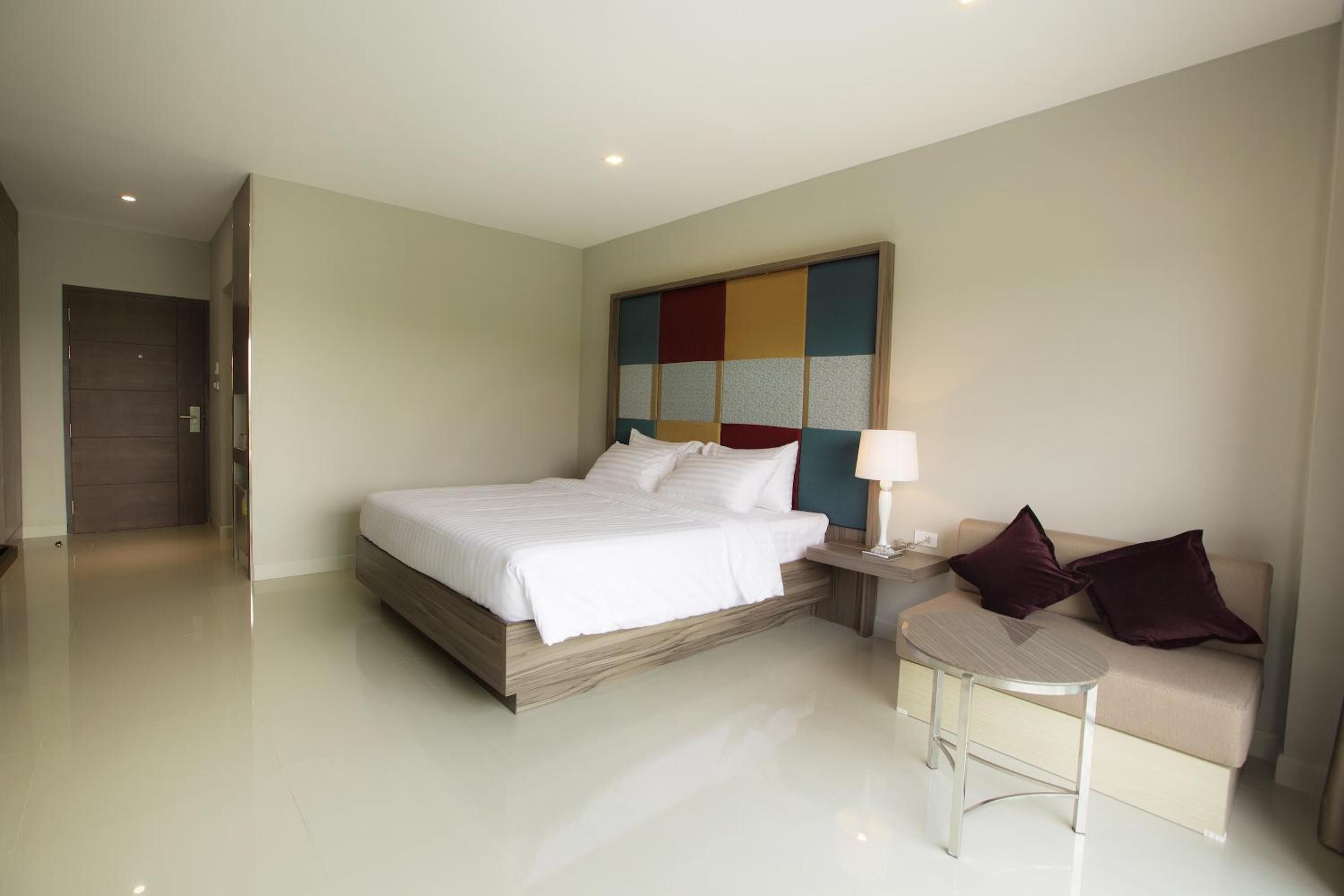 Una habitación de hotel moderna con una cama grande, una mesita de noche con lámpara, un sofá y una obra de arte abstracta y colorida en la pared.