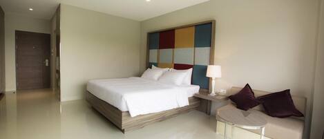 Una habitación de hotel moderna con una cama grande, una mesita de noche con lámpara, un sofá y una obra de arte abstracta y colorida en la pared.