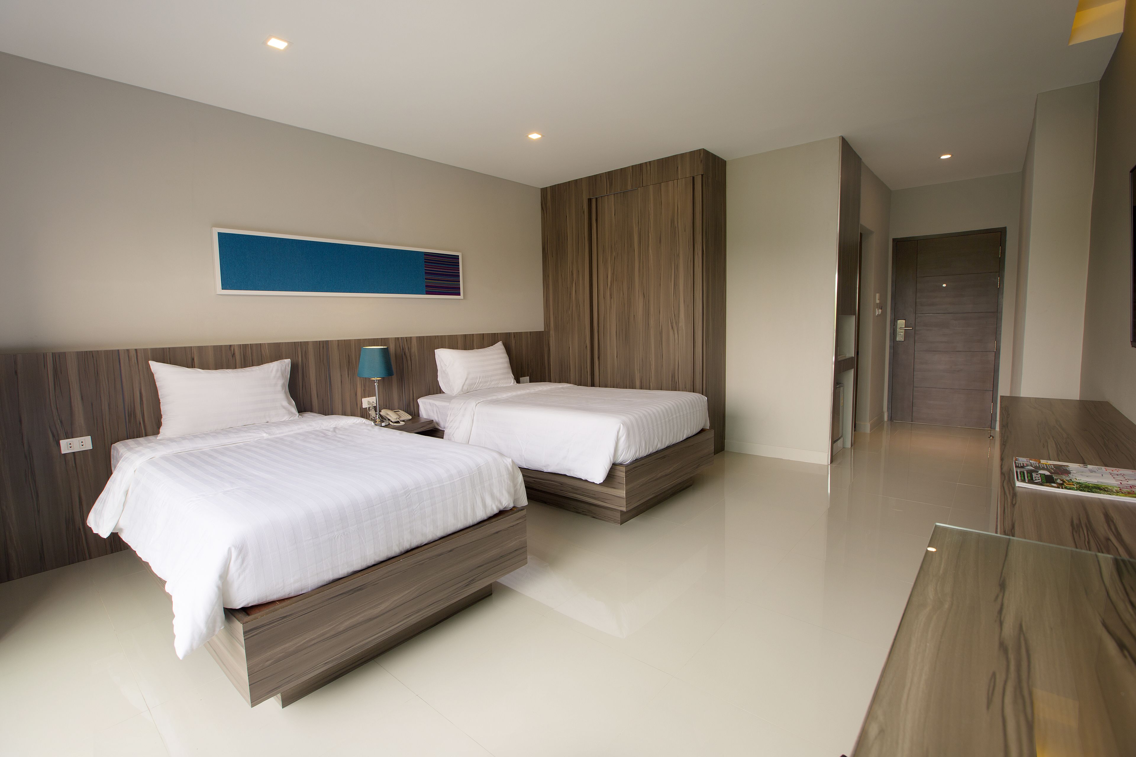 A modern hotel room with two beds, a wooden headboard, a blue accent wall, and a wooden reception desk.