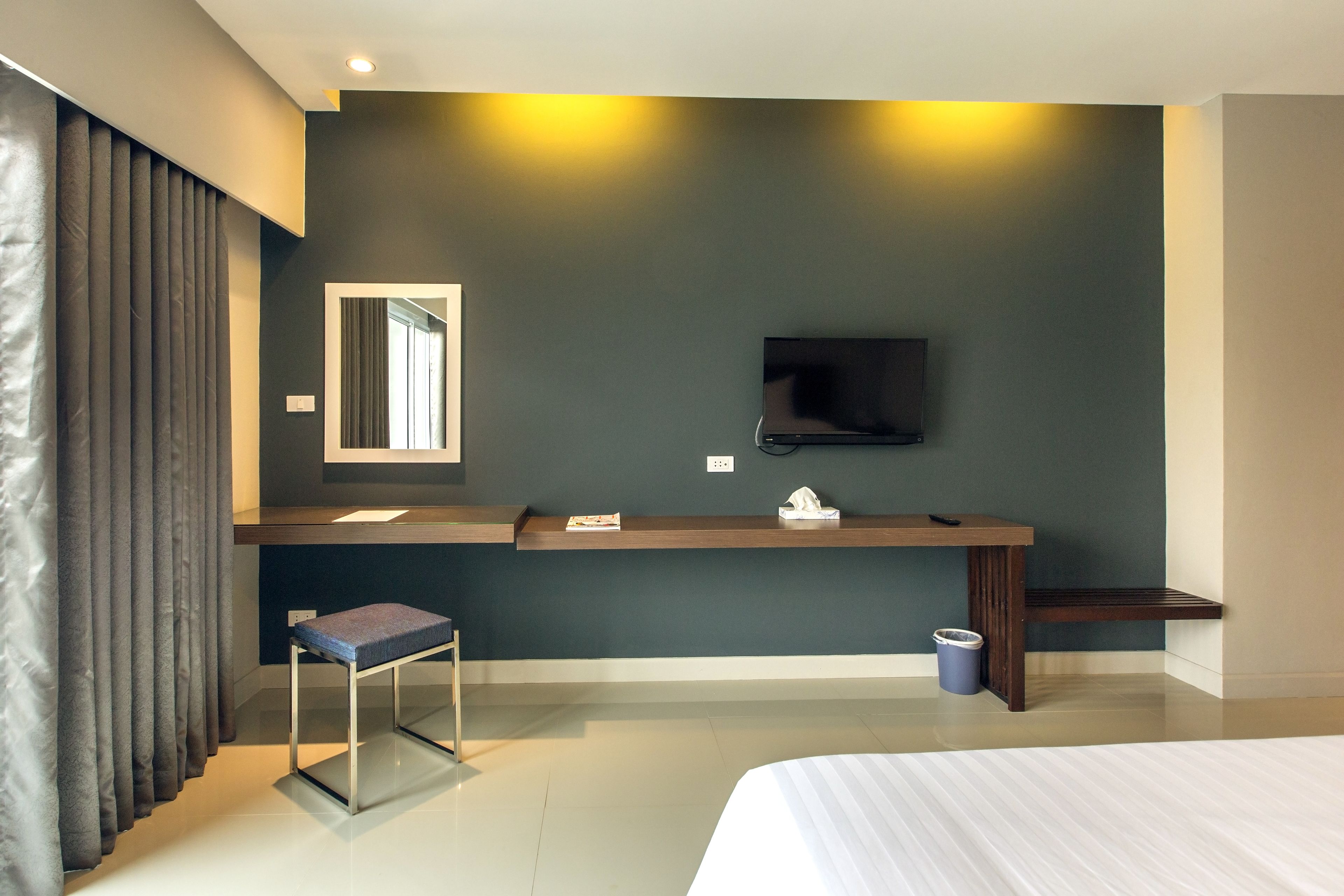 A modern hotel room with a large bed, a bedside table, a small stool, a TV mounted on the wall, and a dark-colored wall.