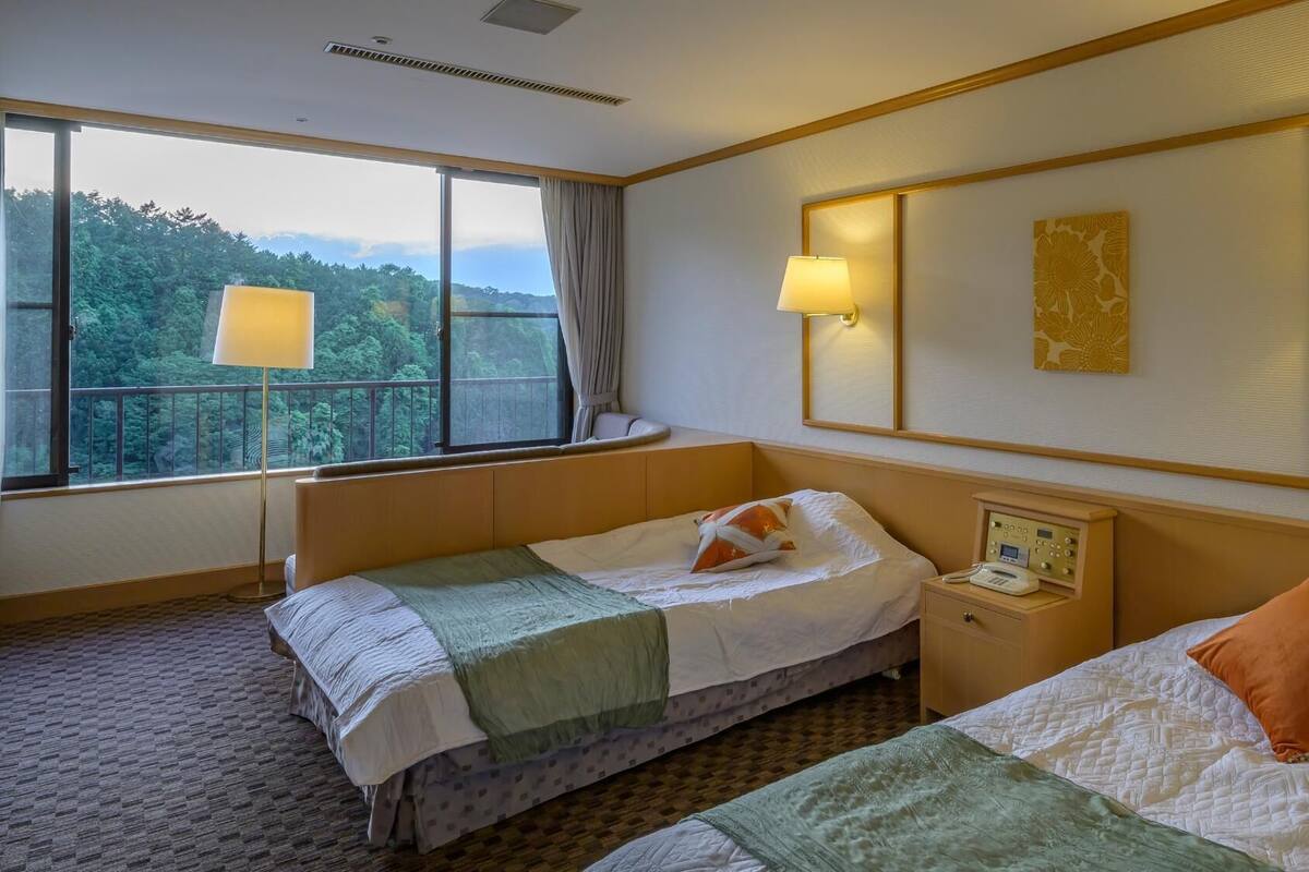 A hotel room with two beds, a bedside table with a phone, a window with a view of trees, and wall-mounted lamps.