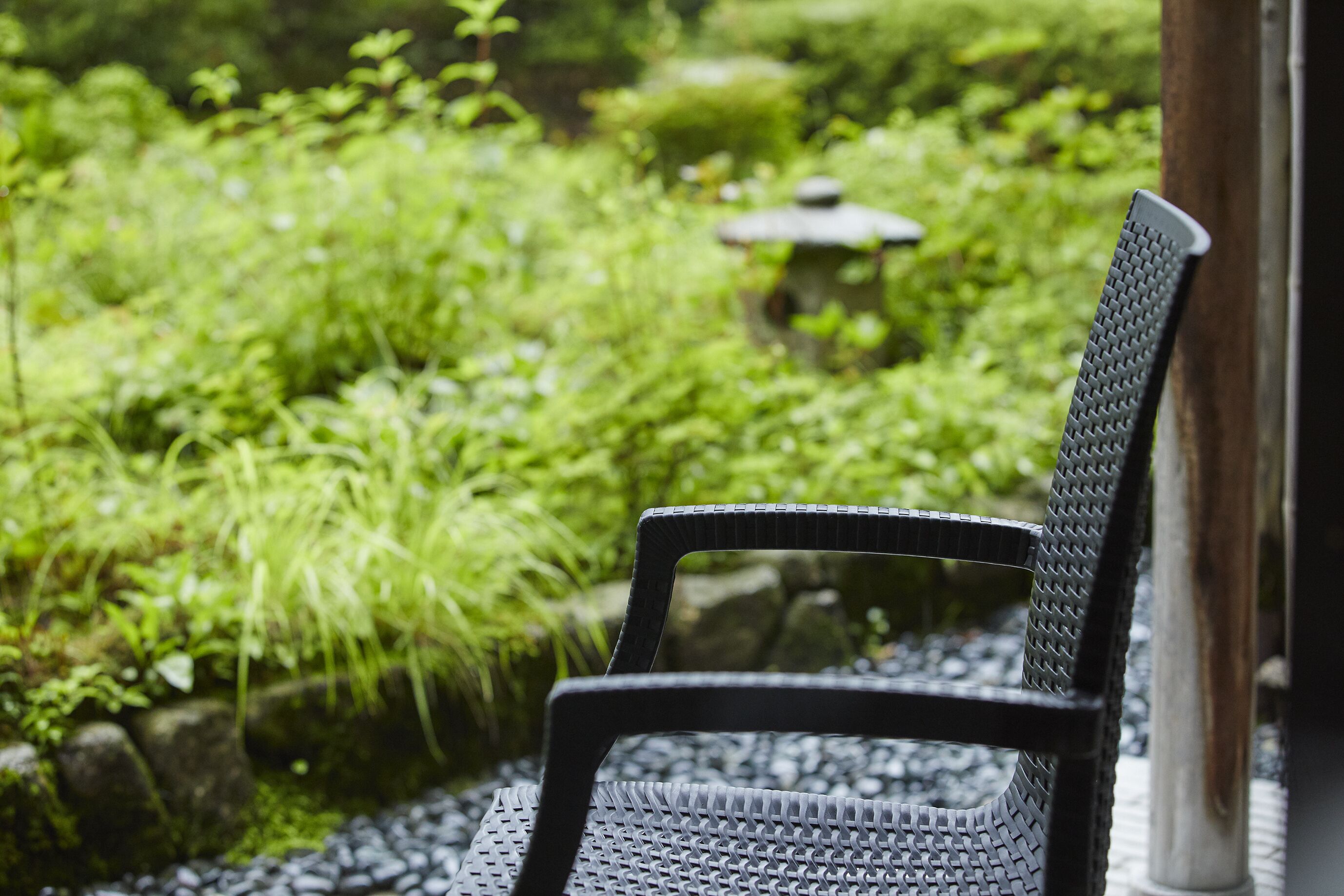 A chair with a textured backrest and seat, positioned outdoors with a view of a garden area featuring greenery and a stone structure in the background.