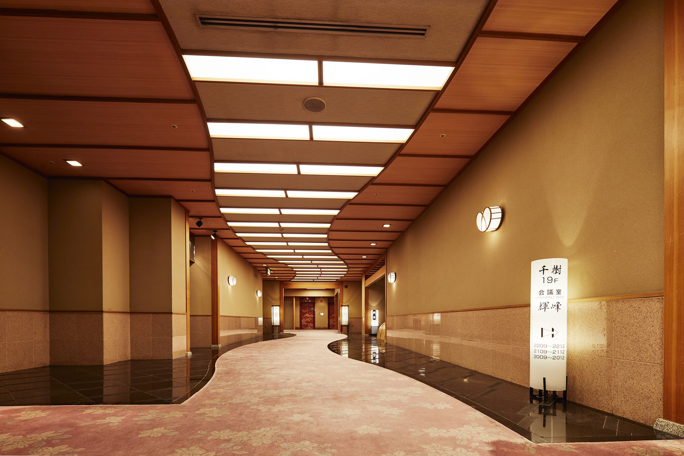 A hotel corridor with a curved carpet, wooden panel ceiling, and a sign indicating room numbers.