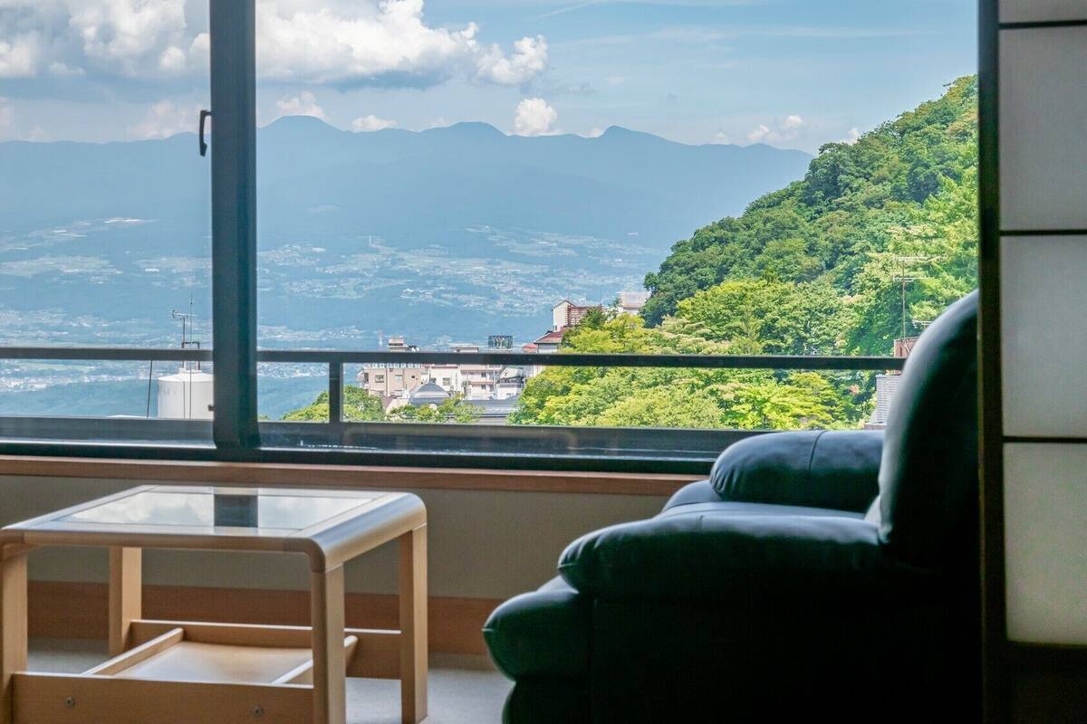 A room with a view of a mountainous landscape, a balcony with a railing, a chair, and a small table.