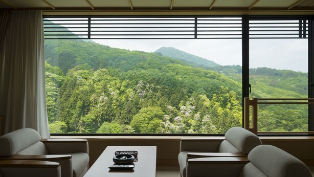 A room with a large window offering a view of a forested mountain. There is a table with a remote control and a decorative bowl. Two sofas are positioned facing the window. - Fukuichi (Shibukawa)