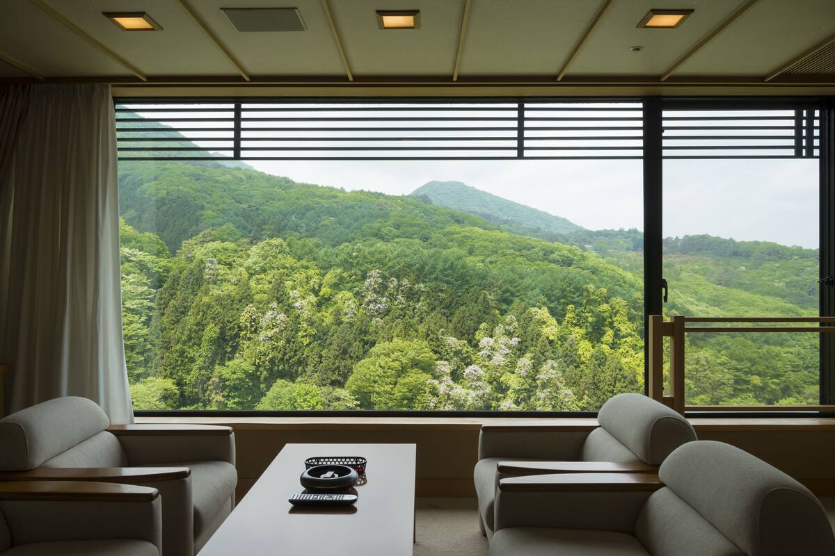 A room with a large window offering a view of a forested mountain. There is a table with a remote control and a decorative bowl. Two sofas are positioned facing the window.