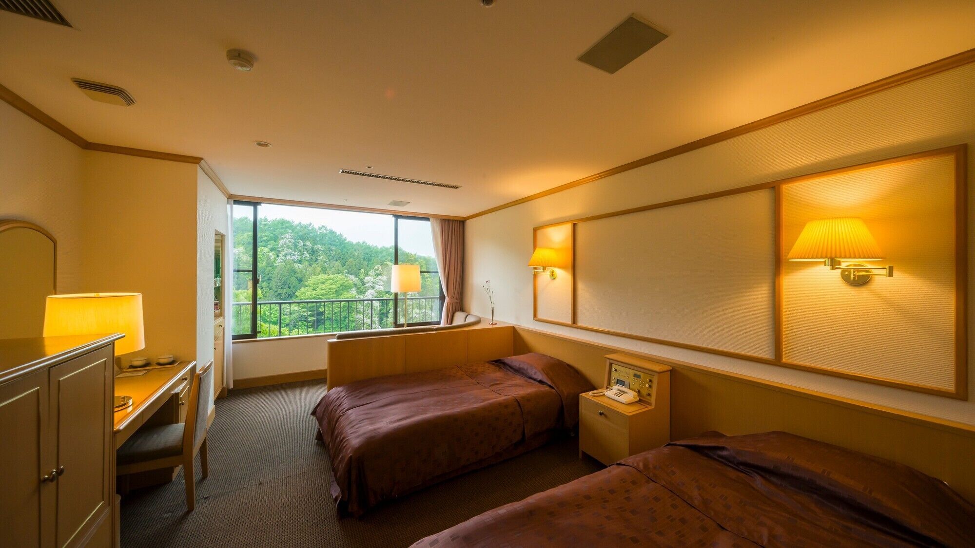 Habitación de hotel con dos camas, un escritorio, una silla, un aparador, una ventana con vista a árboles y una lámpara de pared.