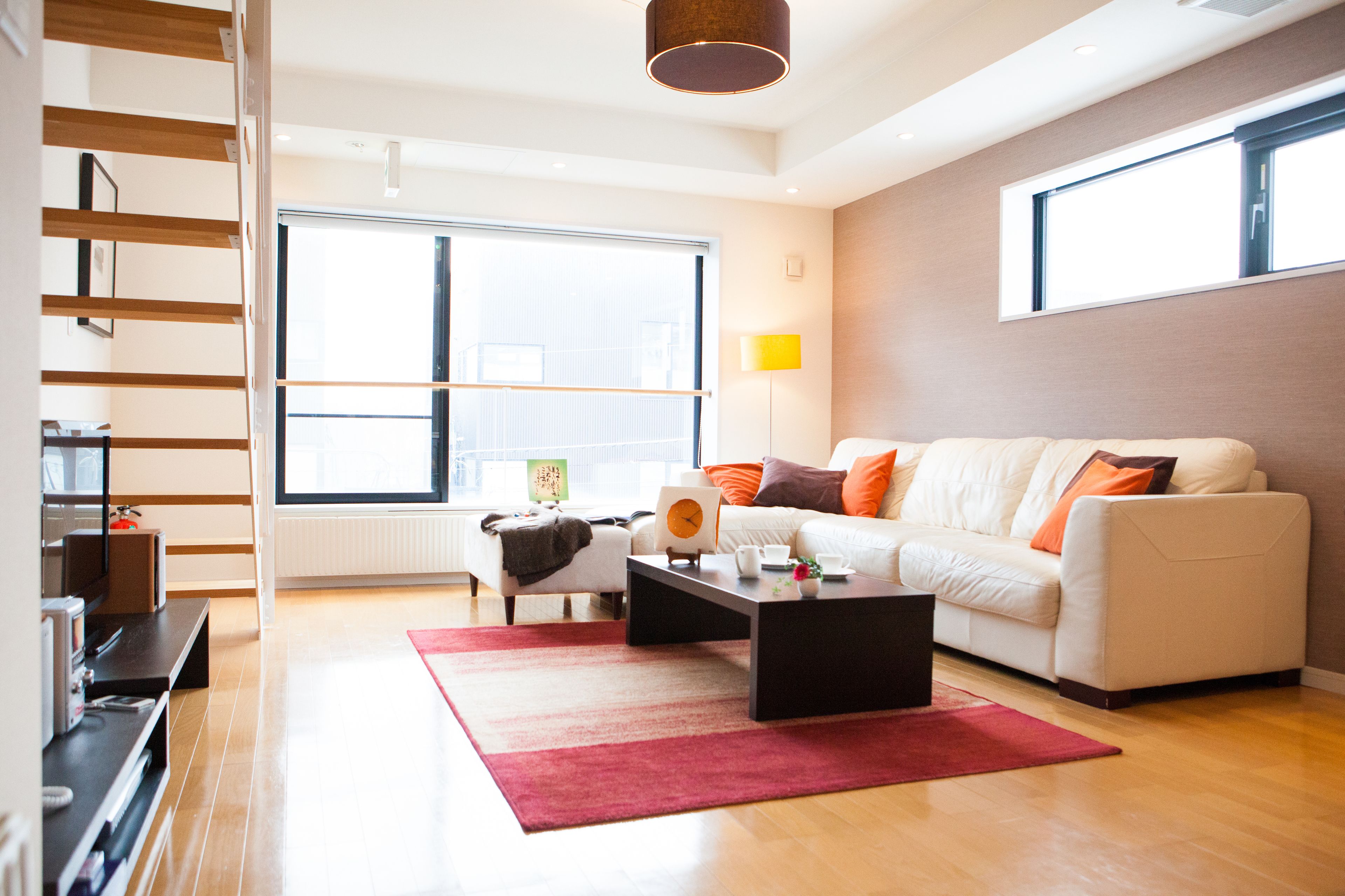 Een moderne woonkamer met een witte bank, een salontafel en een rood vloerkleed. Er is een groot raam dat natuurlijk licht en stadszicht biedt.