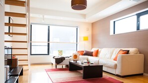 A modern living room with a white sofa, a coffee table, and a red rug. There is a large window providing natural light and a view of the city. - Powder Haven (Kutchan)
