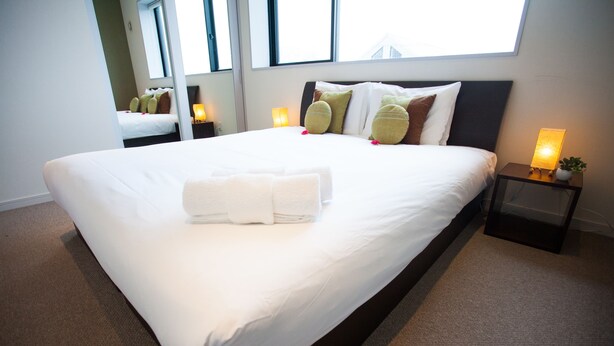 A neatly made bed with white linens and a folded towel, a bedside table with a lamp, and a window with a view of the sky. - Powder Haven (Kutchan)