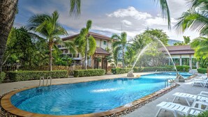 A swimming pool with a water fountain, surrounded by palm trees and a building with a red roof. - Whispering Palms Suite (Koh Samui)