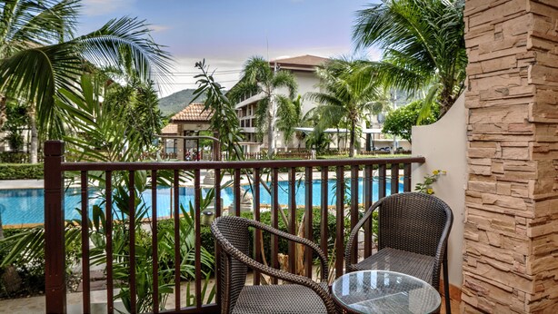 A balcony with wicker chairs and a glass table overlooking a swimming pool and a resort building. - Whispering Palms Suite (Koh Samui)