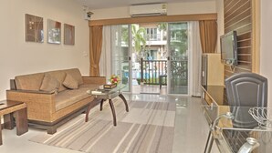A modern living room with a sofa, coffee table, and a TV. There is a balcony with a view of a pool and palm trees. - Whispering Palms Suite (Koh Samui)