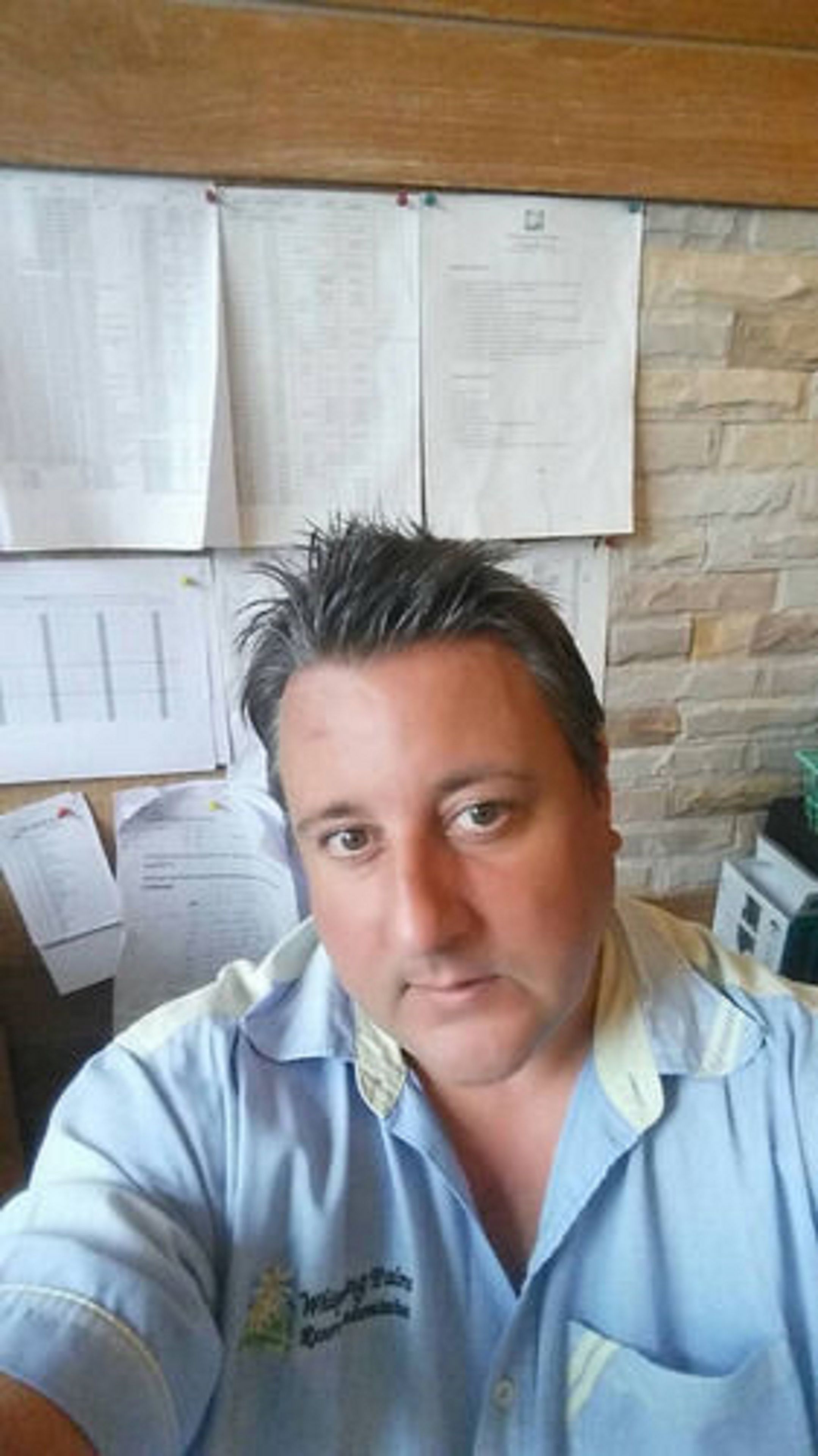 Un hombre con una camisa azul claro con un logo, sentado en una habitación con pared de piedra y papeles sujetos a ella.