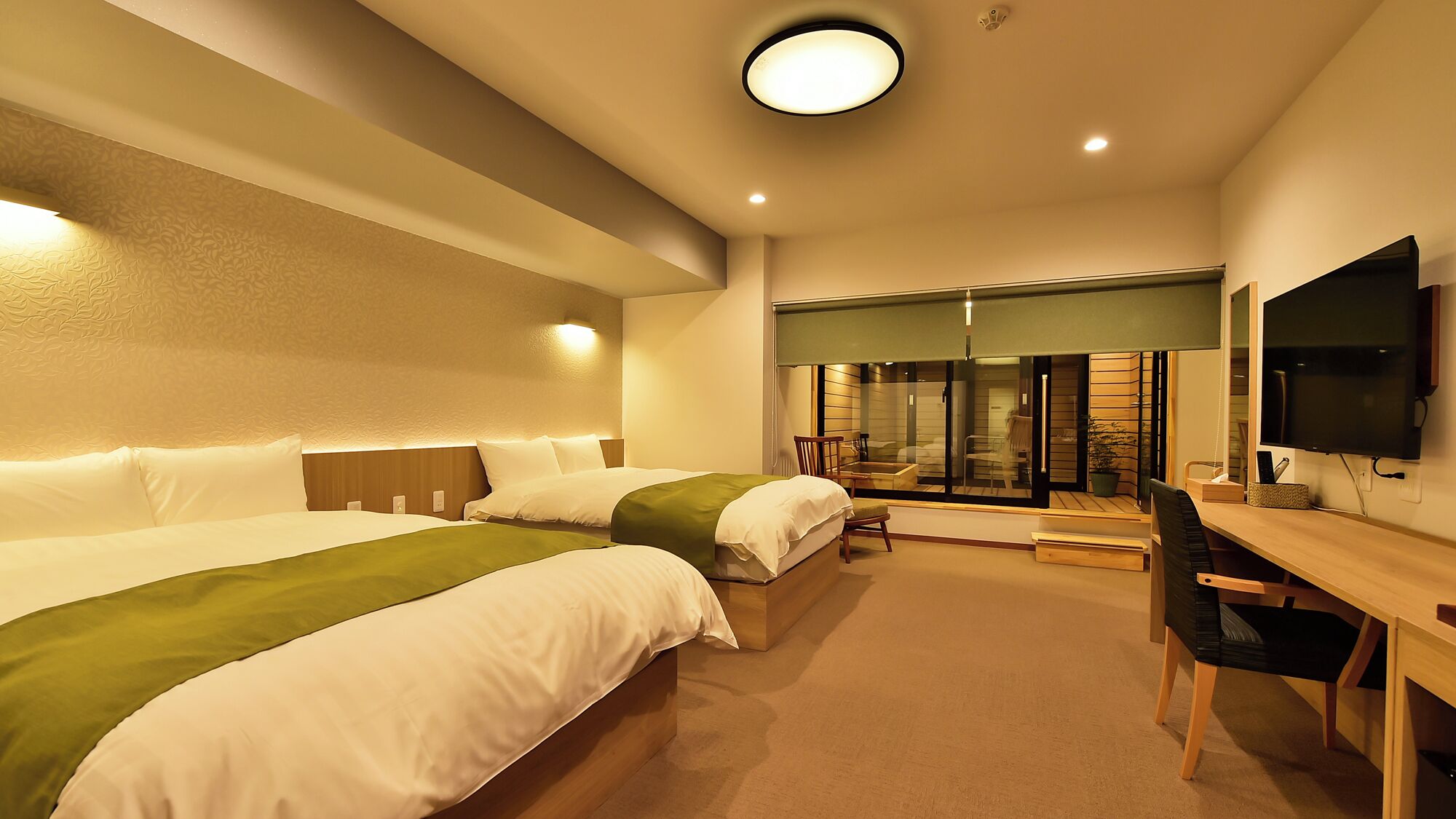 A hotel room with two beds, a desk, a chair, a TV, and a sliding door leading to a balcony.