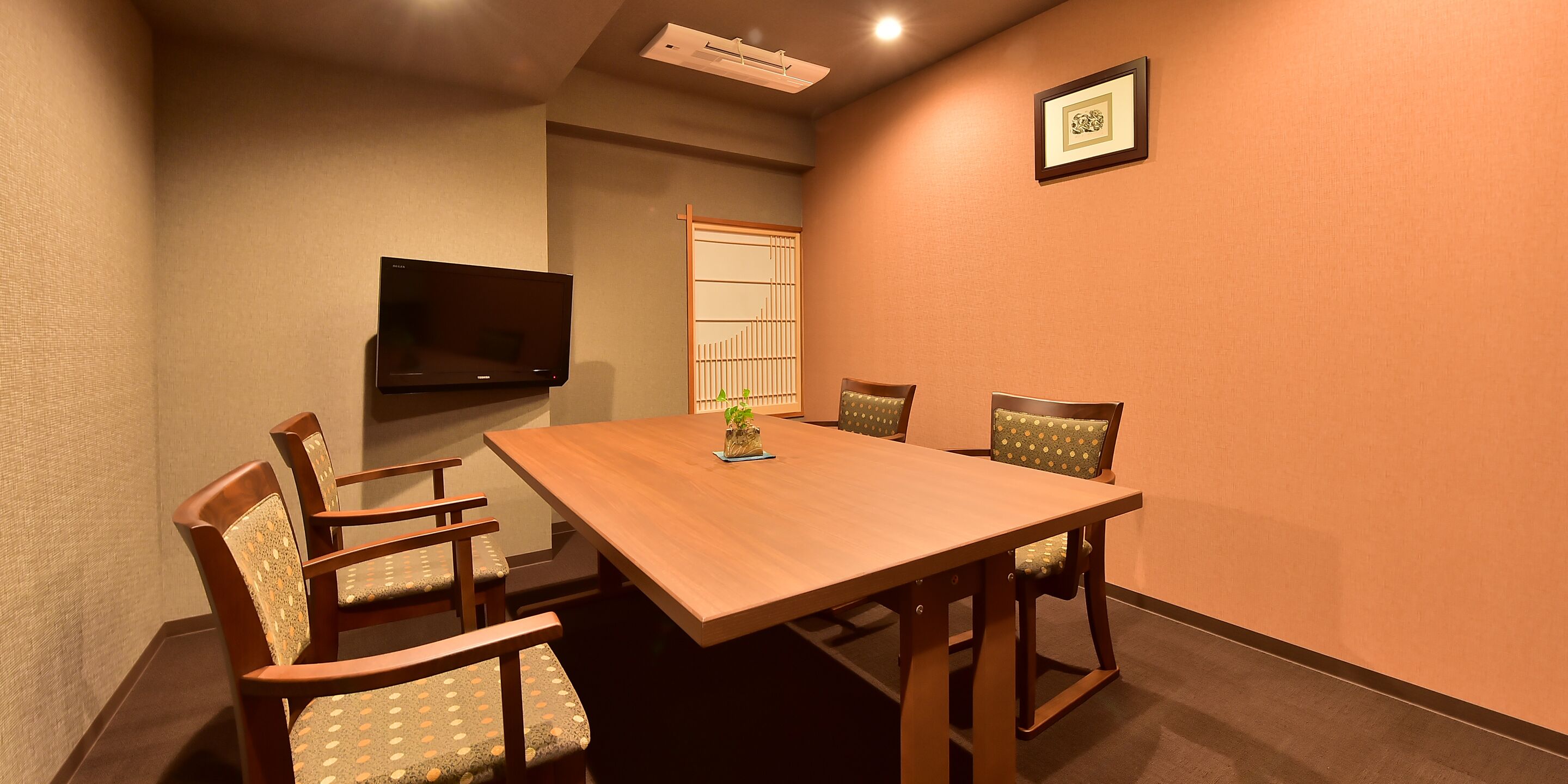A conference room with a wooden table, four chairs, a television mounted on the wall, and a framed picture.