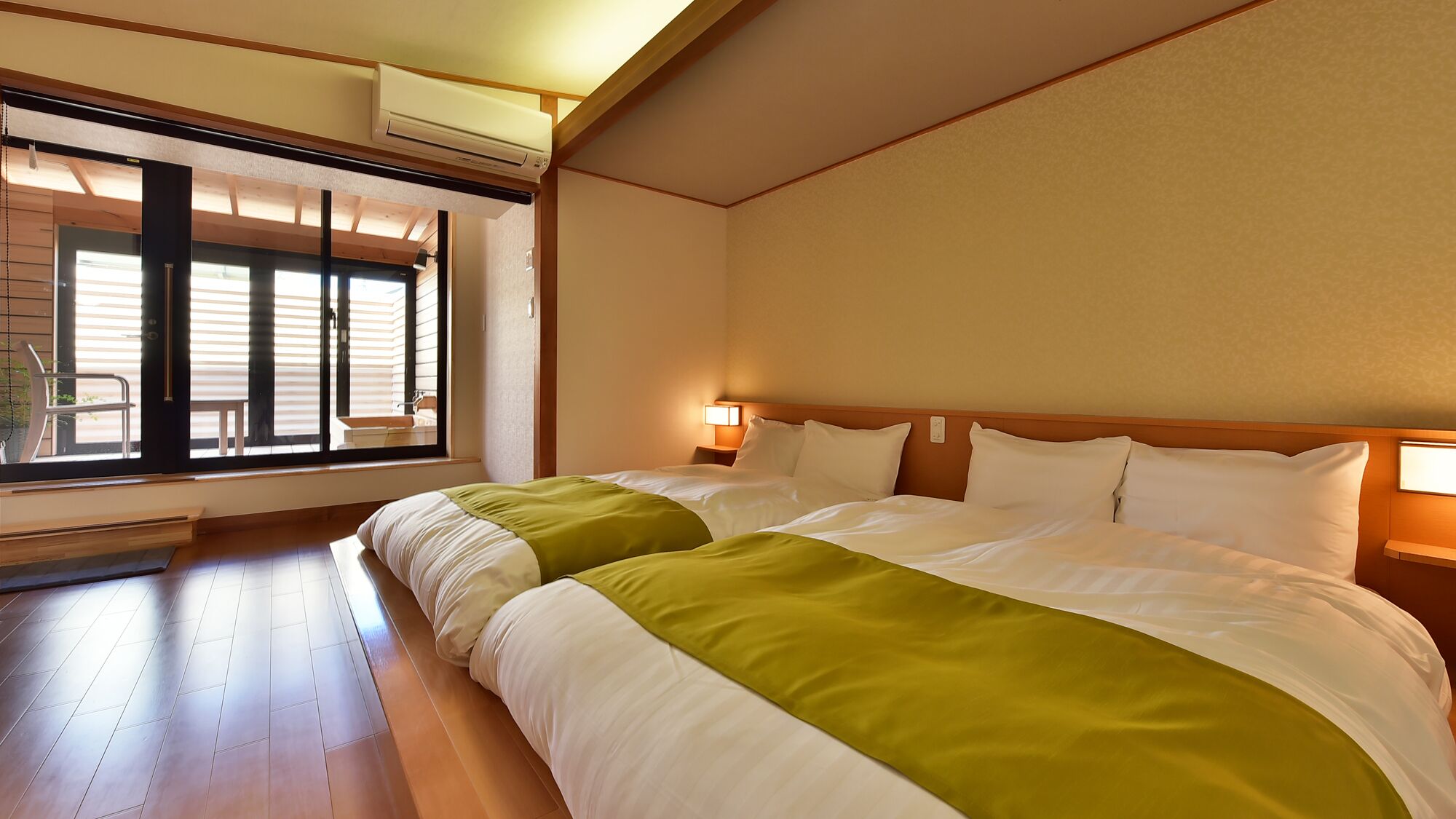 A hotel room with two beds, a window with blinds, an air conditioning unit, and a bedside lamp.