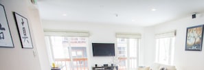 A spacious living room with a large flat-screen TV mounted on the wall, a sofa, and a coffee table. There are two windows with curtains, and a balcony is visible through the windows.
