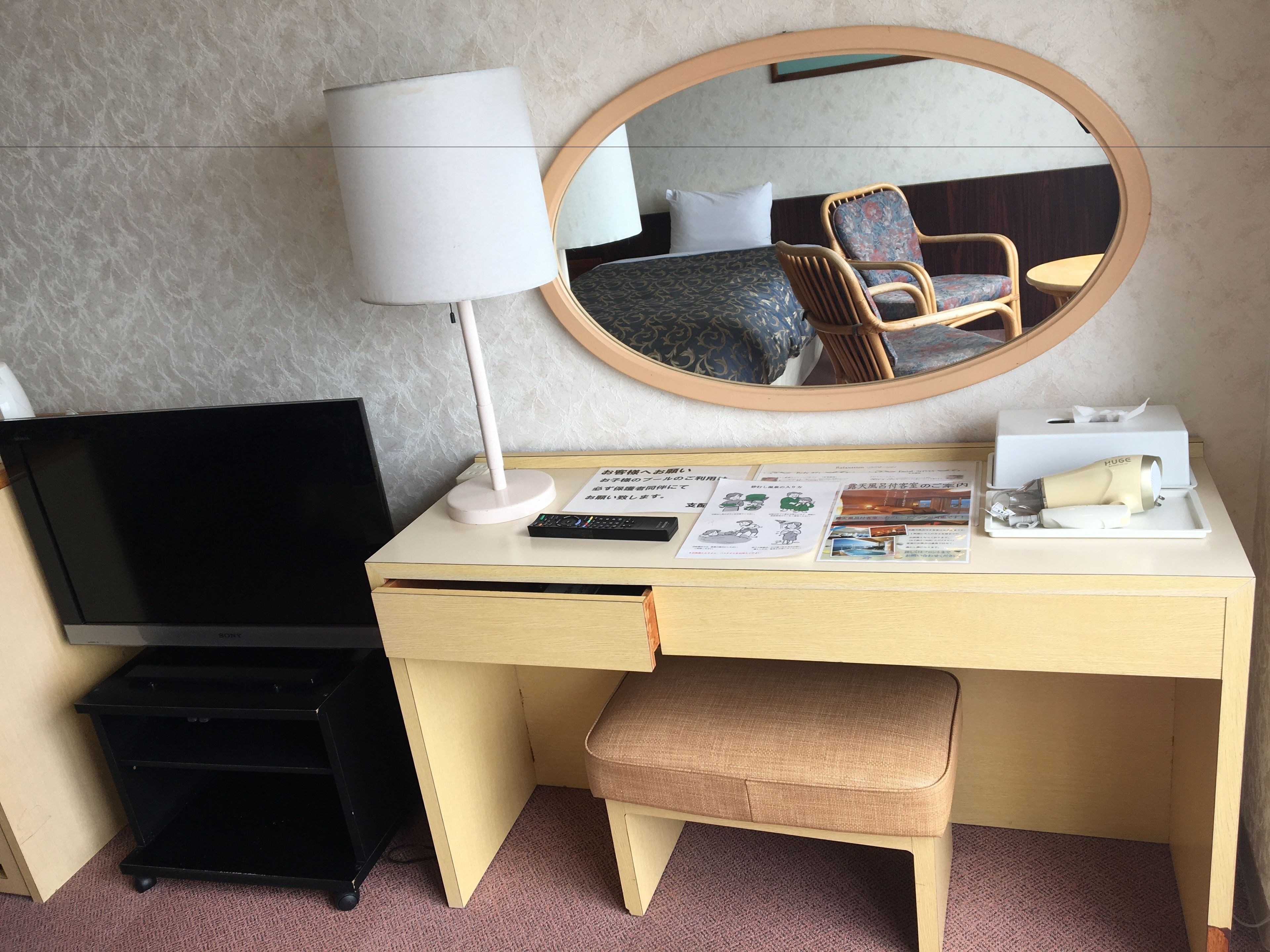 A hotel room with a desk, a television, a lamp, a mirror, a chair, a small table, and a bench.