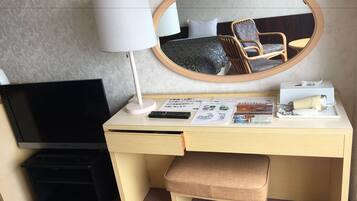 A hotel room with a desk, a television, a lamp, a mirror, a chair, a small table, and a bench.
