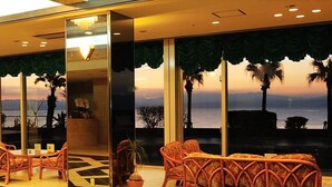 A hotel lobby with wicker furniture, a reception desk, and a large window with a view of the sea and palm trees. - Ibusuki Seaside Hotel (Ibusuki)