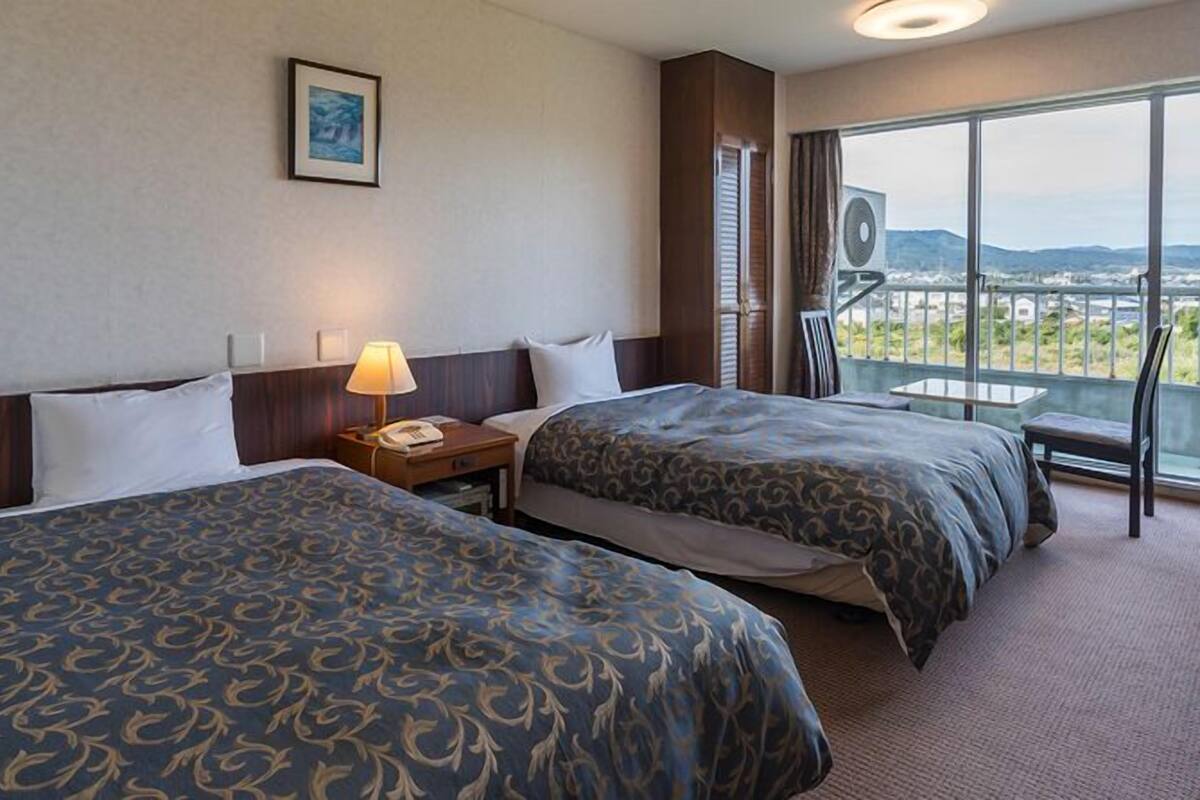 A hotel room with two beds, a nightstand, a lamp, a framed picture on the wall, and a balcony with a view.