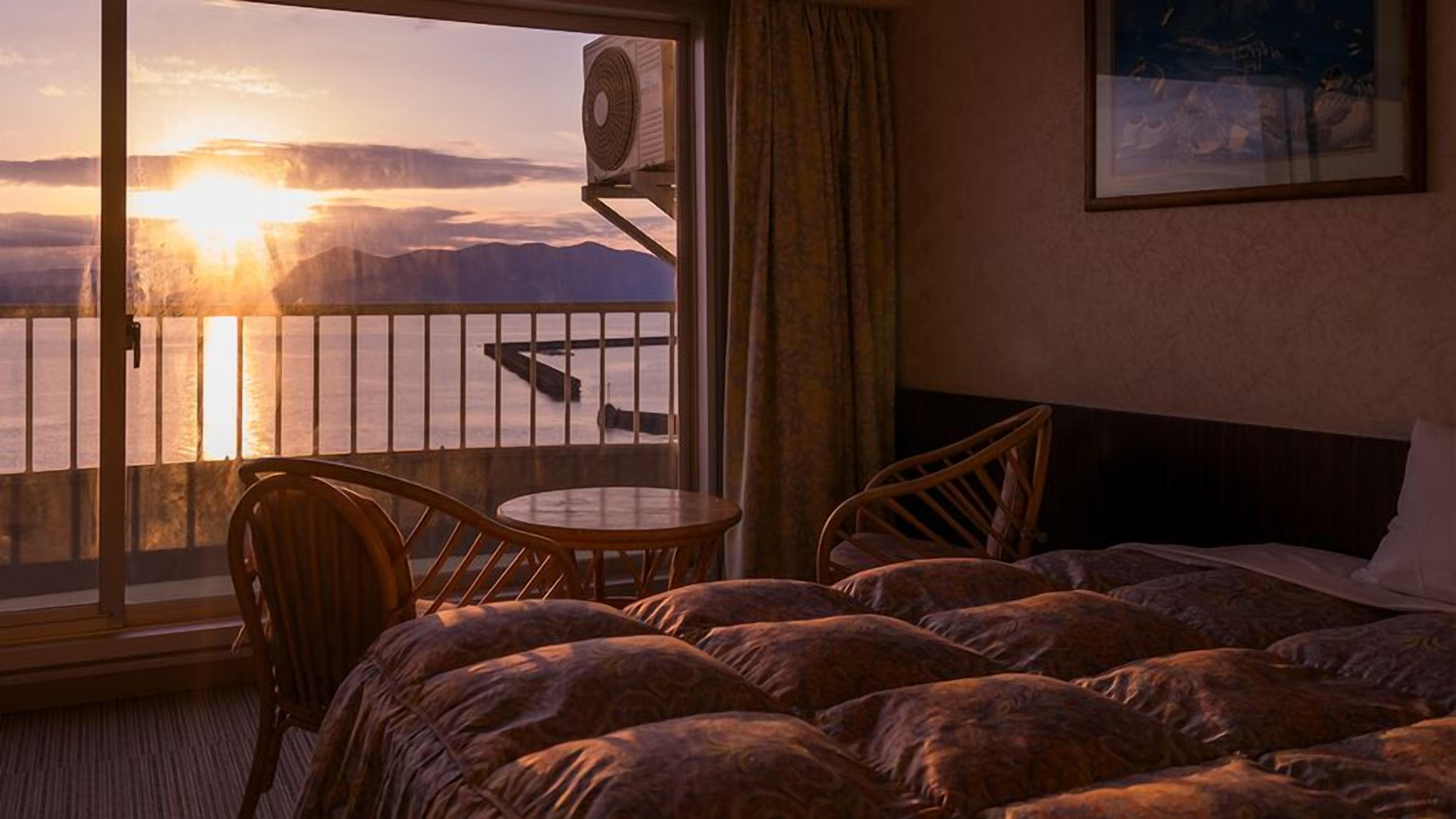 A hotel room with a bed, a chair, a small table, and a balcony with a view of the sunset over the water.
