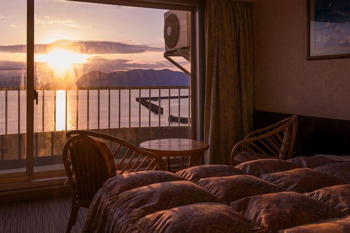 A hotel room with a bed, a chair, a small table, and a balcony with a view of the sunset over the water.