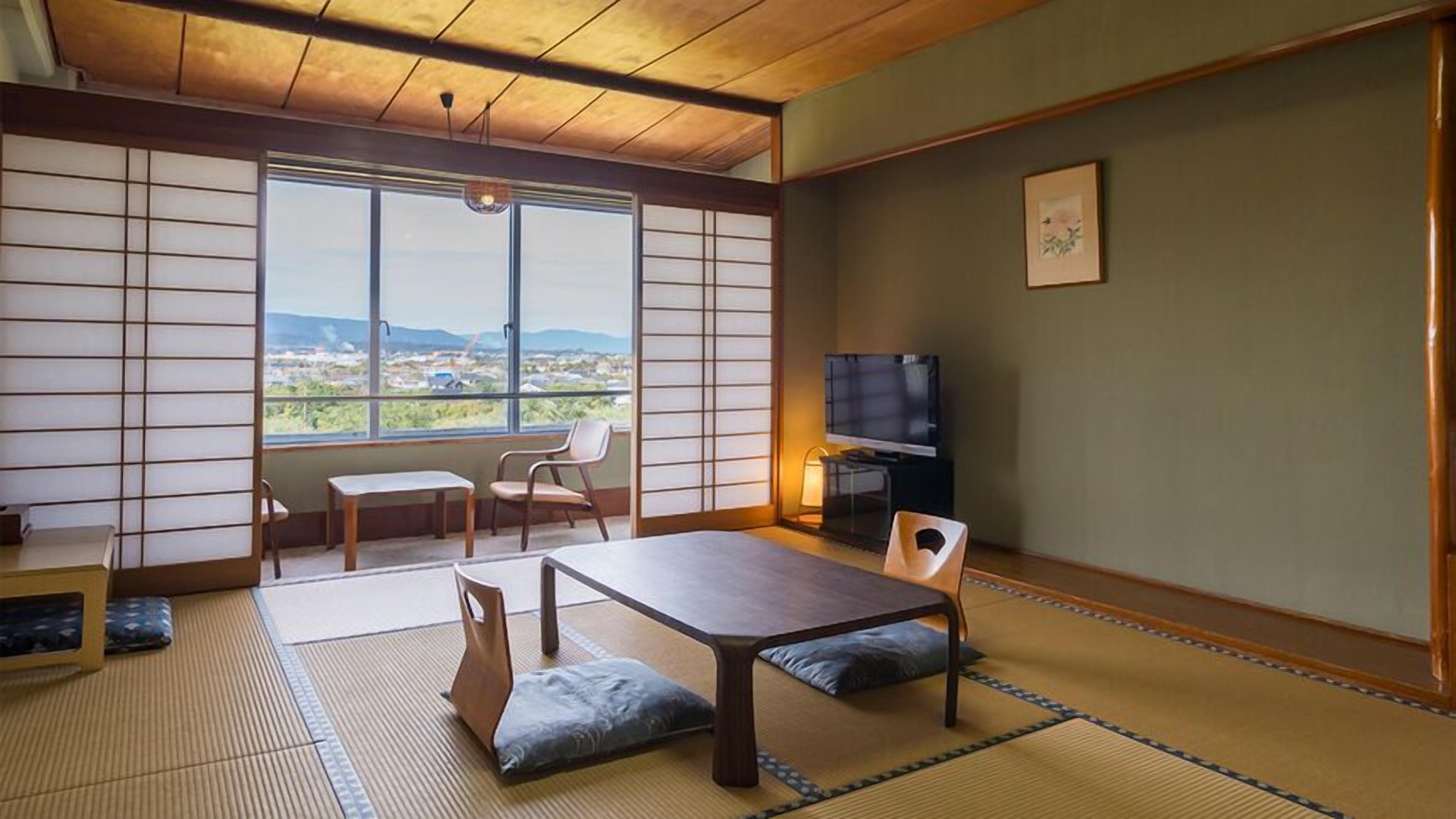 A traditional Japanese room with tatami flooring, a low table, chairs, a television, and a view of the city.
