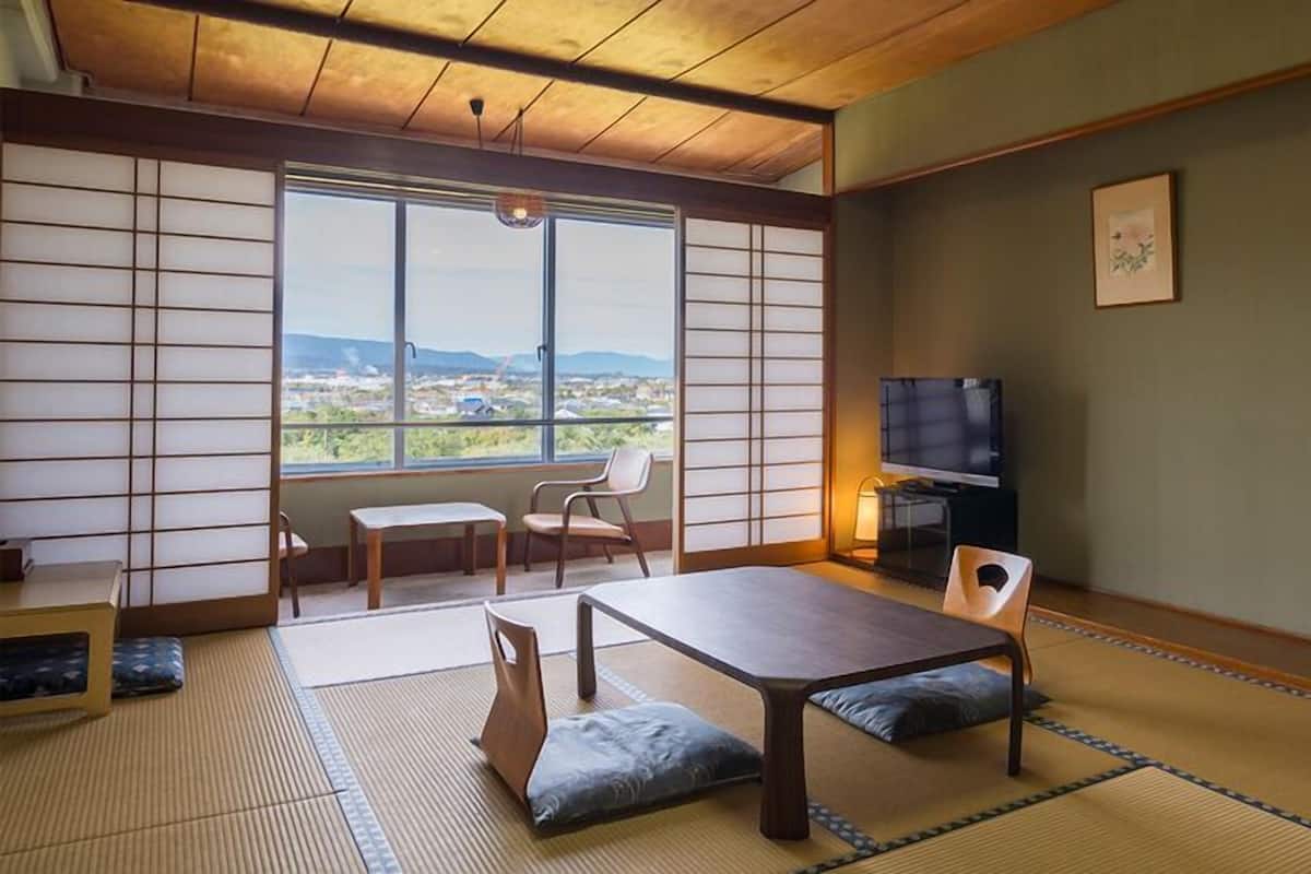 A traditional Japanese room with tatami flooring, a low table, chairs, a television, and a view of the city.