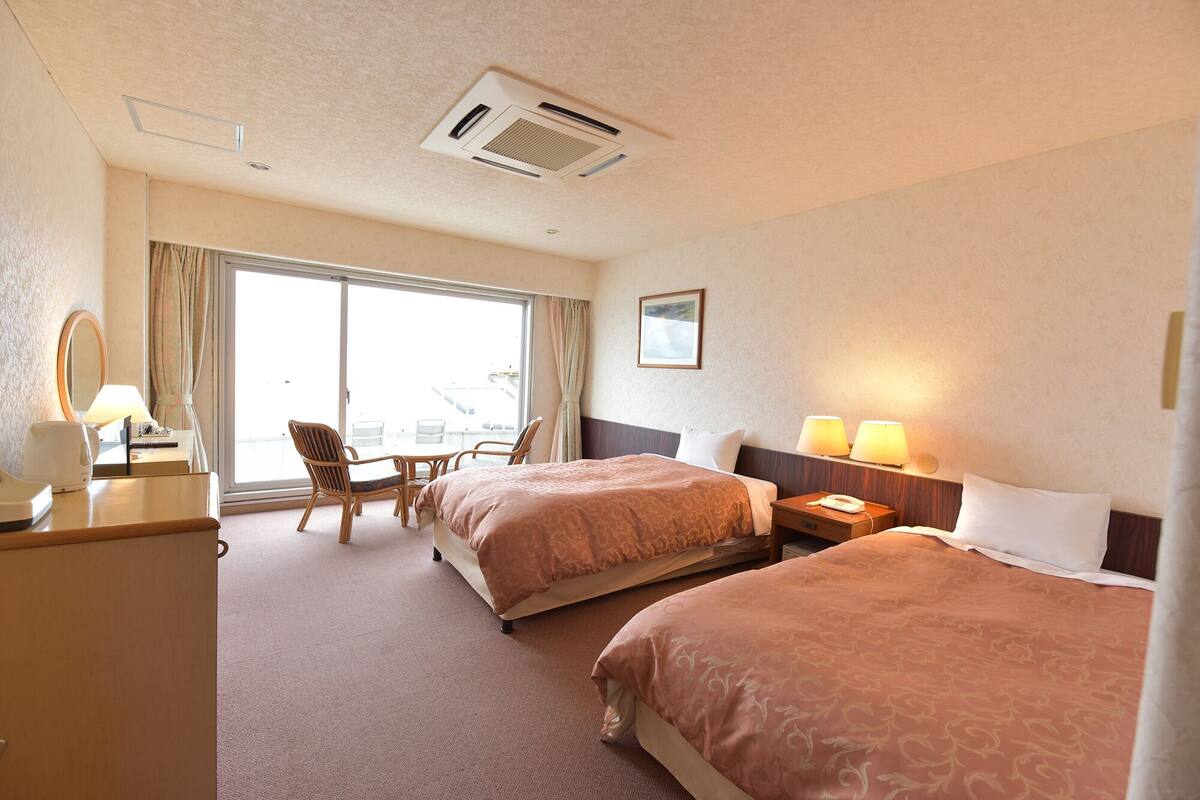 A hotel room with two beds, a desk with a lamp, a chair, and a large window.